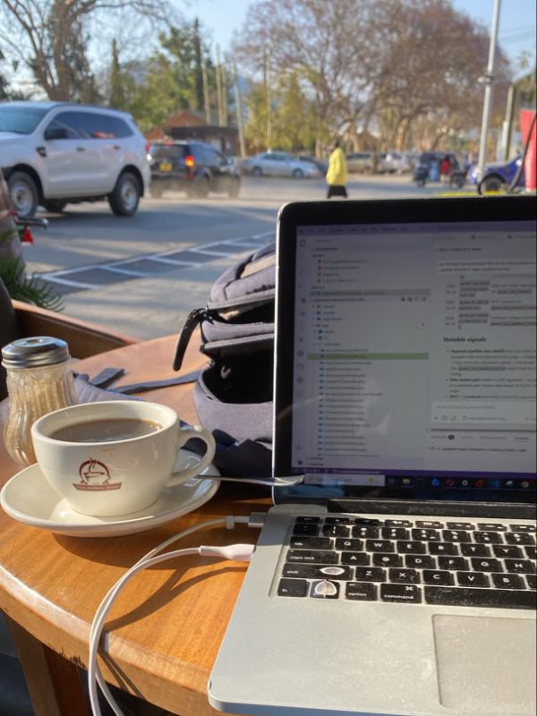 A laptop open on a café table, code on screen, a street and hills behind it.